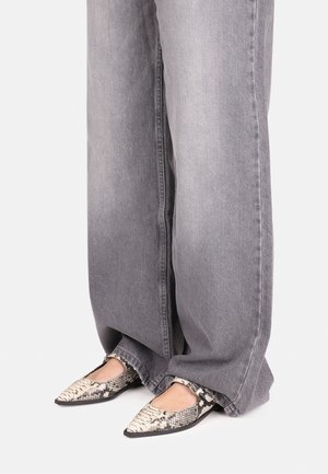 Person trägt weite graue Jeans und flache Schuhe mit spitzer Spitze und Schlangenprint und steht vor weißem Hintergrund.