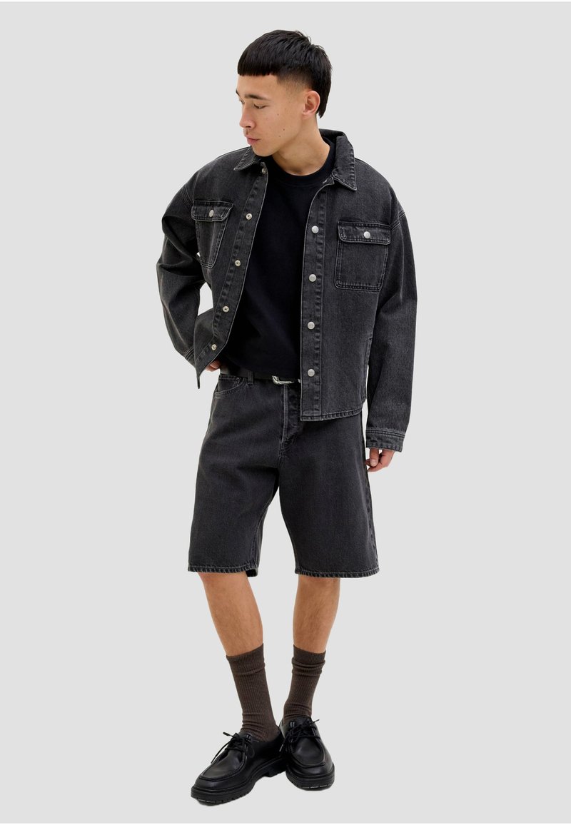 Jeune homme portant une veste en jean noire, un T-shirt noir, un short en jean noir, des chaussettes marron et des chaussures noires, debout avec une main sur la hanche.