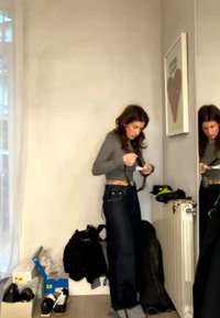 Γυναίκα με γκρι μπλούζα μακριού μανικιού και σκούρα τζιν προσαρμόζει μια ζώνη σε ένα δωμάτιο με παπούτσια, ρούχα και μεγάλο καθρέφτη στον τοίχο.