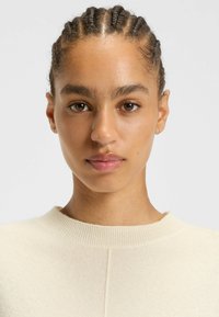 Heller Cremepulli mit rundem Ausschnitt und gerippten Akzenten, der eine glatte Textur aufweist. Haare in glatten Zöpfen nach hinten frisiert, mit minimalem Schmuck.