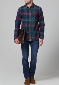 Camisa de franela a cuadros en colores azul marino, verde y burdeos, diseño abotonado; combinada con jeans oscuros y un bolso mensajero de cuero marrón.