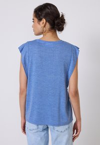 Top azul sin mangas de tejido suave con un ajuste relajado. Con pliegues en los hombros y un escote redondeado, combinado con jeans azul claro.
