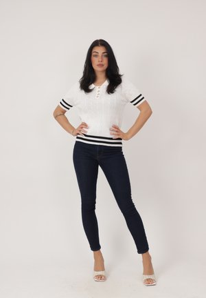 Donna con lunghi capelli scuri, in piedi con le mani sui fianchi, indossa una maglia bianca a maniche corte a righe nere e jeans skinny scuri, tacchi bianchi.