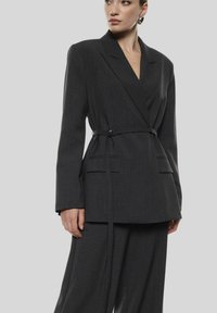 Donna che indossa un blazer cinturato grigio scuro e pantaloni a gamba larga abbinati, in piedi su uno sfondo grigio chiaro uniforme.