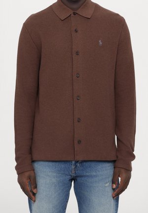 Mężczyzna w brązowej koszuli polo na guziki z długim rękawem i ciemnymi guzikami oraz jasnoniebieskich dżinsach, stojący na jednolitym tle.