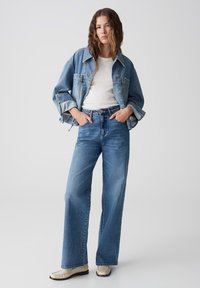 Denimjack met een relaxte pasvorm, lichtblauwe kleur, gecombineerd met high-waisted wijde jeans in een iets donkerder blauw. Crèmekleurige schoenen.