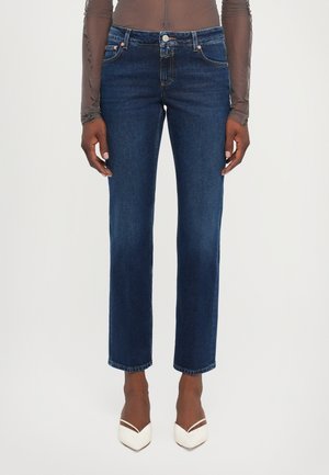 Mørkeblå denimjeans med rett ben-design, fem lommer, knappelukking og subtile sømdetaljer, kombinert med hvite høyhælte sko.