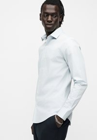 Άνδρας με σκουρόχρωμη επιδερμίδα και dreadlocks που φορά ανοιχτό μπλε πουκάμισο με κουμπιά και σκούρο παντελόνι, κοιτάζει στο πλάι με το ένα χέρι στην τσέπη.