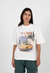 Kvinne med langt mørkt hår som har på seg en hvit, oversized T-skjorte med et trykk av en vintage gul bil og teksten "In a Rush Racing Club Berlin".