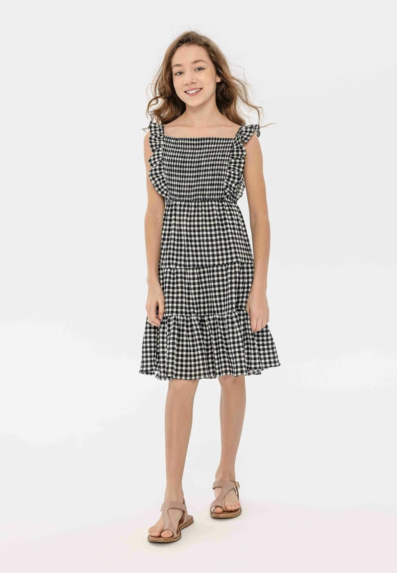 Robe à carreaux noir et blanc avec des manches en volant et un ourlet en couches, fabriquée en tissu léger, portée avec des sandales beige.
