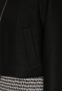 Schwarze Jacke aus strukturiertem Stoff mit einem Reißverschluss und einem aufgenähten Taschen-Detail. Kombiniert mit einer gemusterten Hose in hellen und dunklen Tönen.