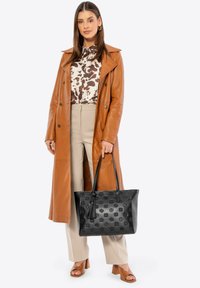 Bolso tote de cuero negro con grabado floral, con un adorno de borlas. Combinado con un trench coat marrón sobre una blusa estampada y pantalones beige.