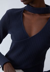 Navy ribbad topp med djup V-ringning och hög krage. Tyget är texturerat och har en åtsittande silhuett och långa ärmar för ökad täckning.