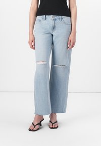 Šviesiai mėlyni nusidėvėję plačių kojų džinsai su matomais kelių plyšiais, klasikiniu penkių kišenių dizainu ir šiek tiek nusidėvėjusiu apvadu. Dėvimi su juodais šlepetėmis.