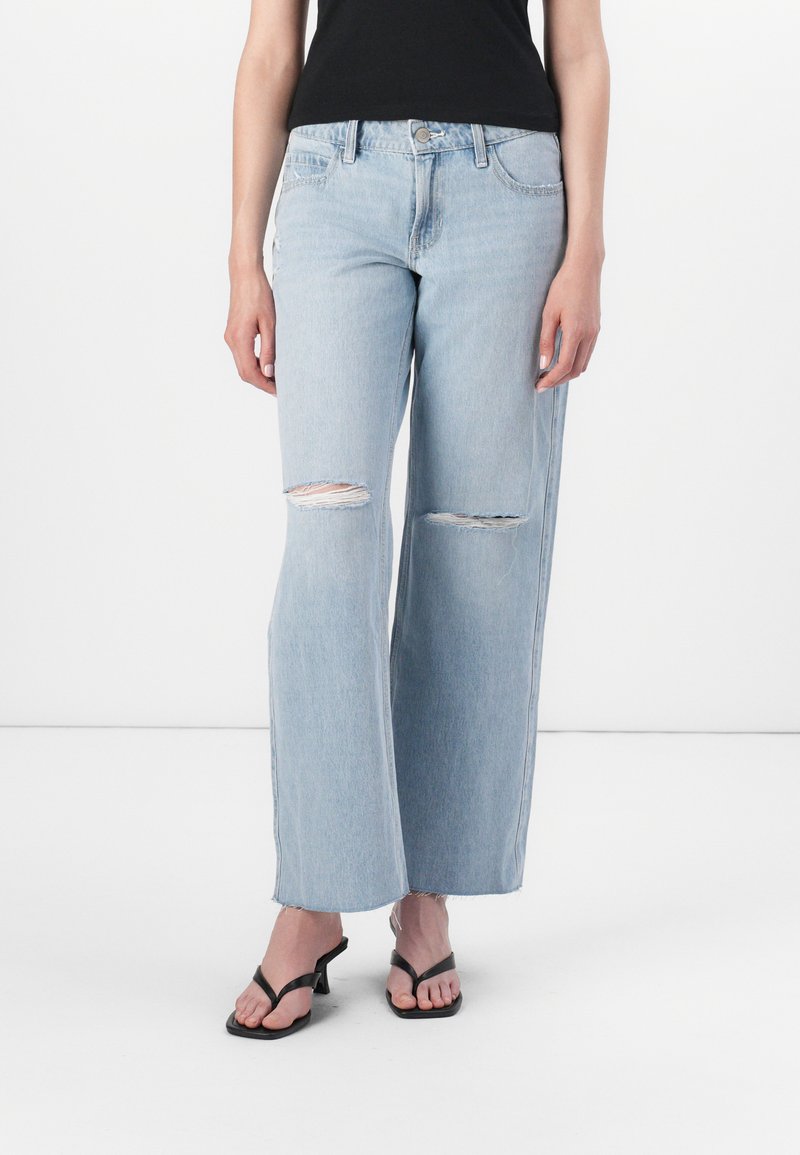 Šviesiai mėlyni nusidėvėję plačių kojų džinsai su matomais kelių plyšiais, klasikiniu penkių kišenių dizainu ir šiek tiek nusidėvėjusiu apvadu. Dėvimi su juodais šlepetėmis.