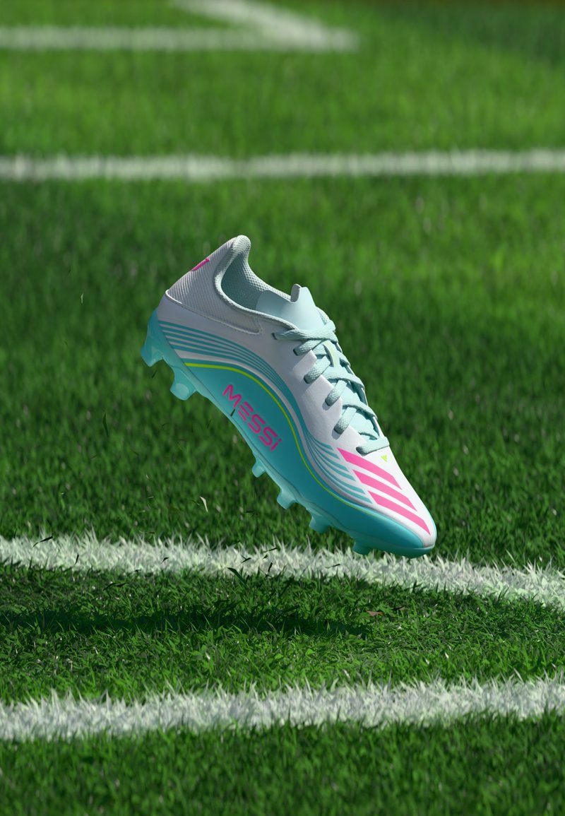 Chaussure de football blanche et turquoise avec des touches roses et le texte « MESSI » suspendu au-dessus d’un terrain en gazon avec des lignes blanches.