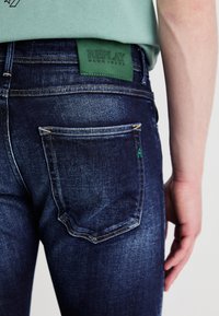 Donkerblauwe denim jeans met lichte stiksels, met een groene leren patch met het label "REPLAY" boven de achterzak. Close-up zijkant weergave.
