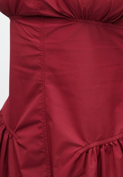 Φόρεμα σε βουργουνδί αποχρώσεις από λείο ύφασμα, με σφηκοφωλιά στη μέση, βολάν στο τελείωμα και αδιάκοπη πλαϊνή ραφή με ελαφριά γυαλάδα.