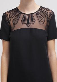 Černé šaty s průhledným vyšívaným výstřihem s geometrickými vzory a krátkými rukávy. Hladká látka pod zdobenou částí.