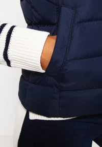 Ręka w kieszeni granatowej pikowanej kurtki noszonej na białym swetrze z czarnymi paskami na rękawie.