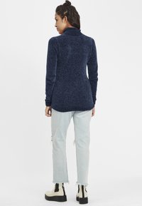 Marineblå, teksturert genser med høy hals og tettsittende design, kombinert med lyseblå slitte jeans og kremfargede ankelstøvler.