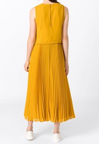 Vestido plisado de color mostaza con diseño sin mangas, que presenta un top ajustado y una falda acampanada. Cierre de cremallera en la parte posterior y textura suave.