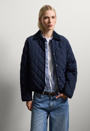 Marineblauwe gestikte jas met een corduroy kraag, voorzien van een knoopsluiting en een textuurpatroon, gedragen over een blauw geruit shirt en spijkerbroeken.