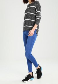 Gestreifter Strickpullover in Schwarz-Weiß, blaue schmale Jeans und schwarze Sportschuhe mit blauen Akzenten. Das Outfit bietet ein lässiges, komfortables Design.