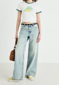 Vit avkortad t-shirt med gröna detaljer och citron grafiskt tryck, ihop med ljusblå vida jeans och gula sneakers. Inkluderar en brun väska.