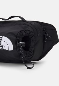 Sac banane noir avec sangle ajustable, poche avant à cordon élastique et logo blanc partiel sur une surface en tissu lisse.