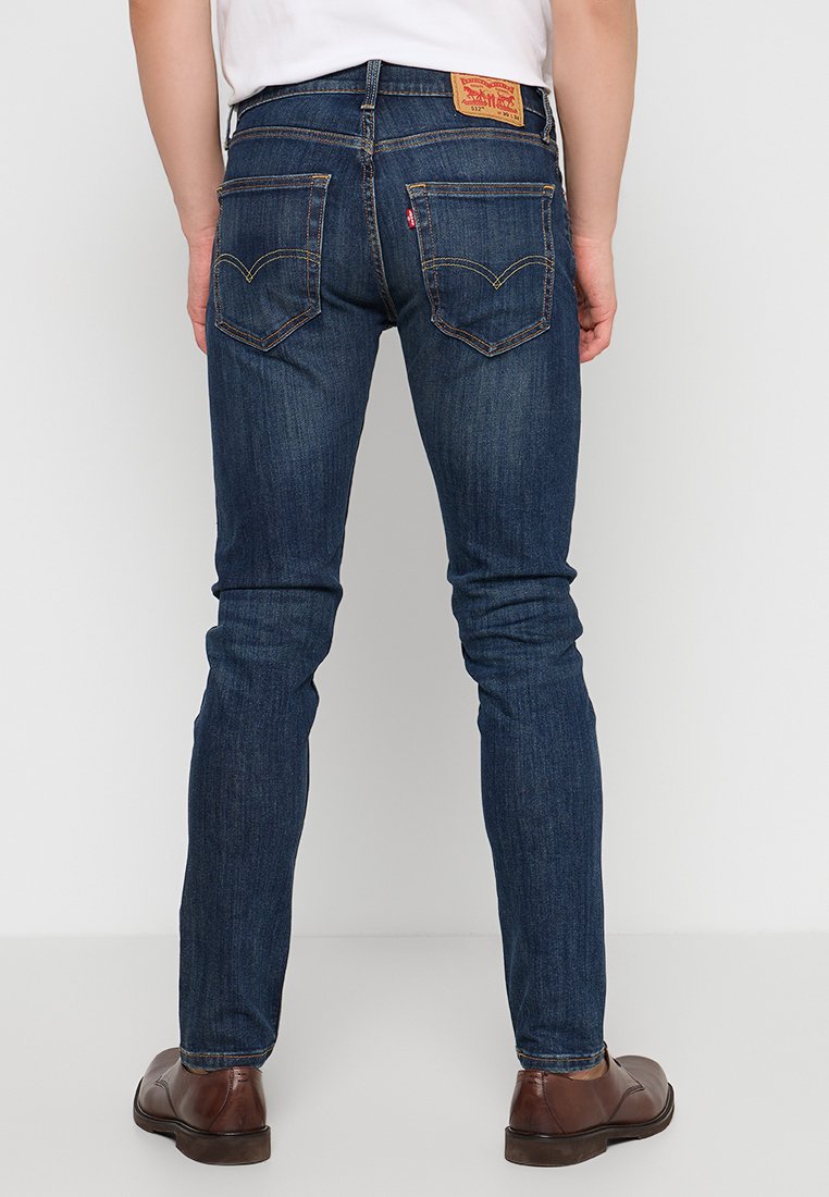 Mężczyzna w ciemnoniebieskich jeansach Levi's o slim fit i brązowych skórzanych butach, stojący tyłem do aparatu na jednolitym tle.