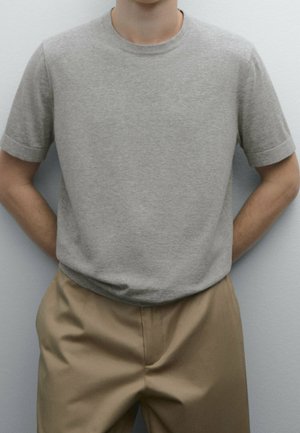 Person standing with hands behind back wearing a plain light gray short-sleeve shirt and beige pants against a neutral background.