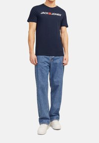 Marineblaue Baumwoll-T-Shirt mit kurzen Ärmeln, ausgestattet mit einem weißen Logo-Druck "JACK & JONES". Kombiniert mit hellblauen geraden Jeans und weißen Sneakern.