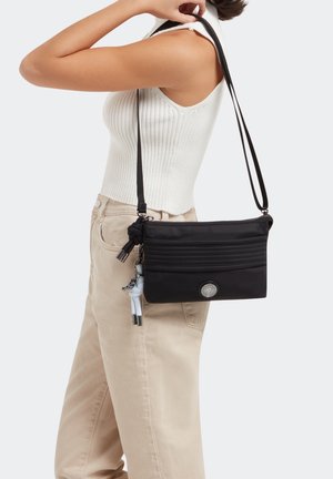Femme portant un haut blanc sans manches et un pantalon beige, avec un sac bandoulière noir orné d'un pompon et d'un porte-clés.