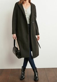 Lang mørkegrøn frakke med hætte og enkelt knaplukning, parret med en lys grøn top, mørke jeans og sorte ankelstøvler. Sort håndtaske inkluderet.