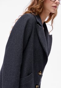 Donkergrijze blazer met een subtiele gestreepte textuur, voorzien van een enkelvoudige knopen, met notch revers en goudkleurige knopen.