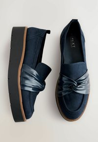 Navy blue slip-on shoes with a suede finish, featuring a twisted synthetic leather strap and a thick rubber sole. Textured design details included.