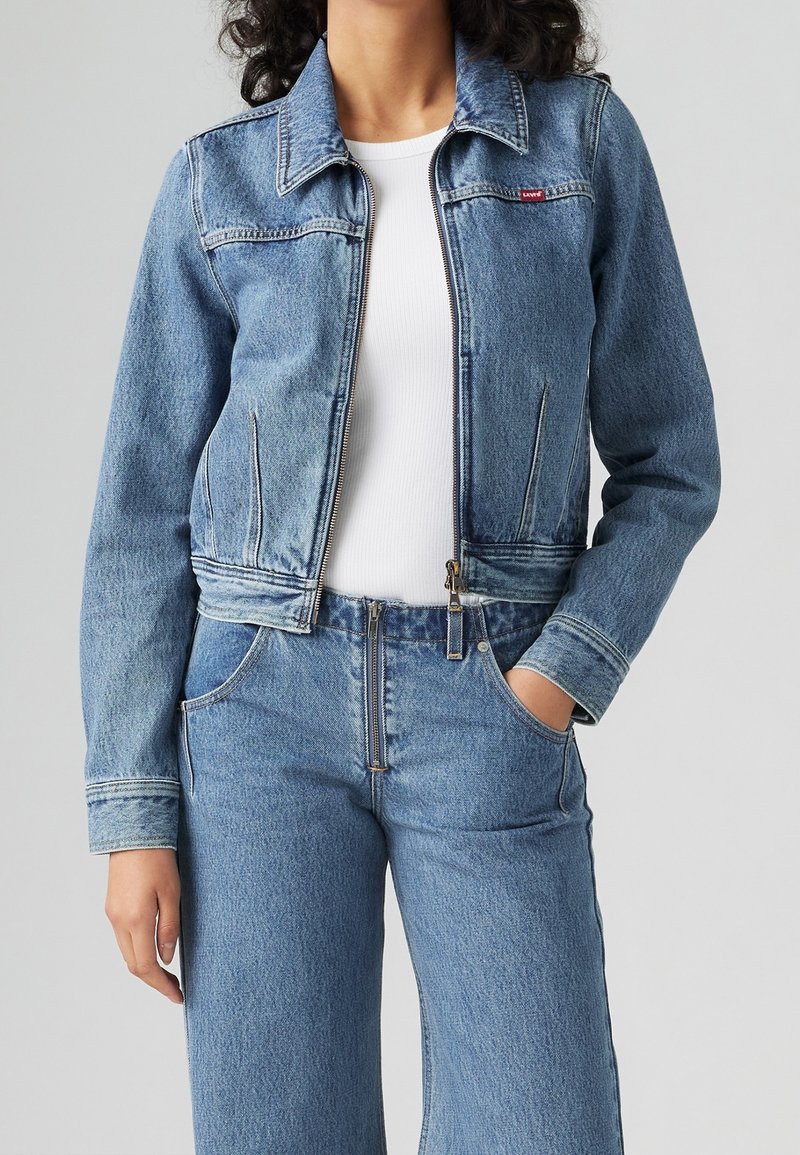 Femme portant une veste en jean bleu clair et un jean taille haute assorti avec un t-shirt blanc côtelé, une main dans la poche, sur fond gris uni.
