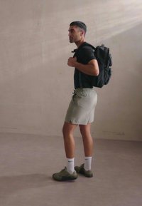 Sac à dos noir, chemise noire à manches courtes, shorts gris clair, chaussettes blanches et chaussures vert olive. Tenue simple et fonctionnelle sur fond neutre.