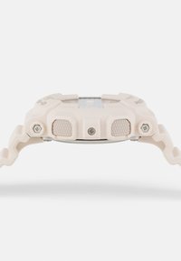 Montre Casio G-Shock en plastique beige clair avec des boutons texturés, un cadran métallique rond et un design de bracelet ondulé unique.