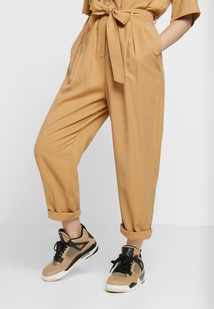 Person iført opkavede beige bukser med et bundet bælte og beige og sorte høje sneakers, stående mod en ensfarvet baggrund.