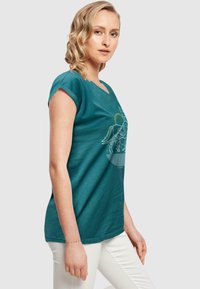 Teal bomulds t-shirt med korte ærmer og v-udskæring, med et hvidt grafisk design af et vinget væsen på forsiden. Båret med hvide bukser.