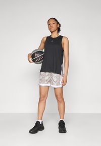 Melns bezpiedeles sporta krekls, šorti ar čūskas rakstu un melnas sporta kurpes. Tur rokās basketbola bumbu ar redzamu logotipu. Balts fons.