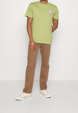 Man staat, draagt een lichtgroen T-shirt met print, bruine jeans met rechte pijpen, witte lage sneakers en een zilveren armband aan de linkerpols.