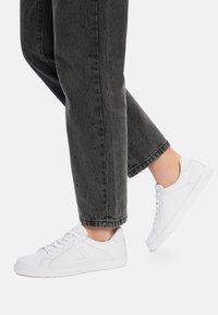 Białe sneakersy z perforowanymi akcentami, sznurowane, z gumową podeszwą, zestawione z przyciętymi czarnymi dżinsami. Prosta i czysta estetyka.