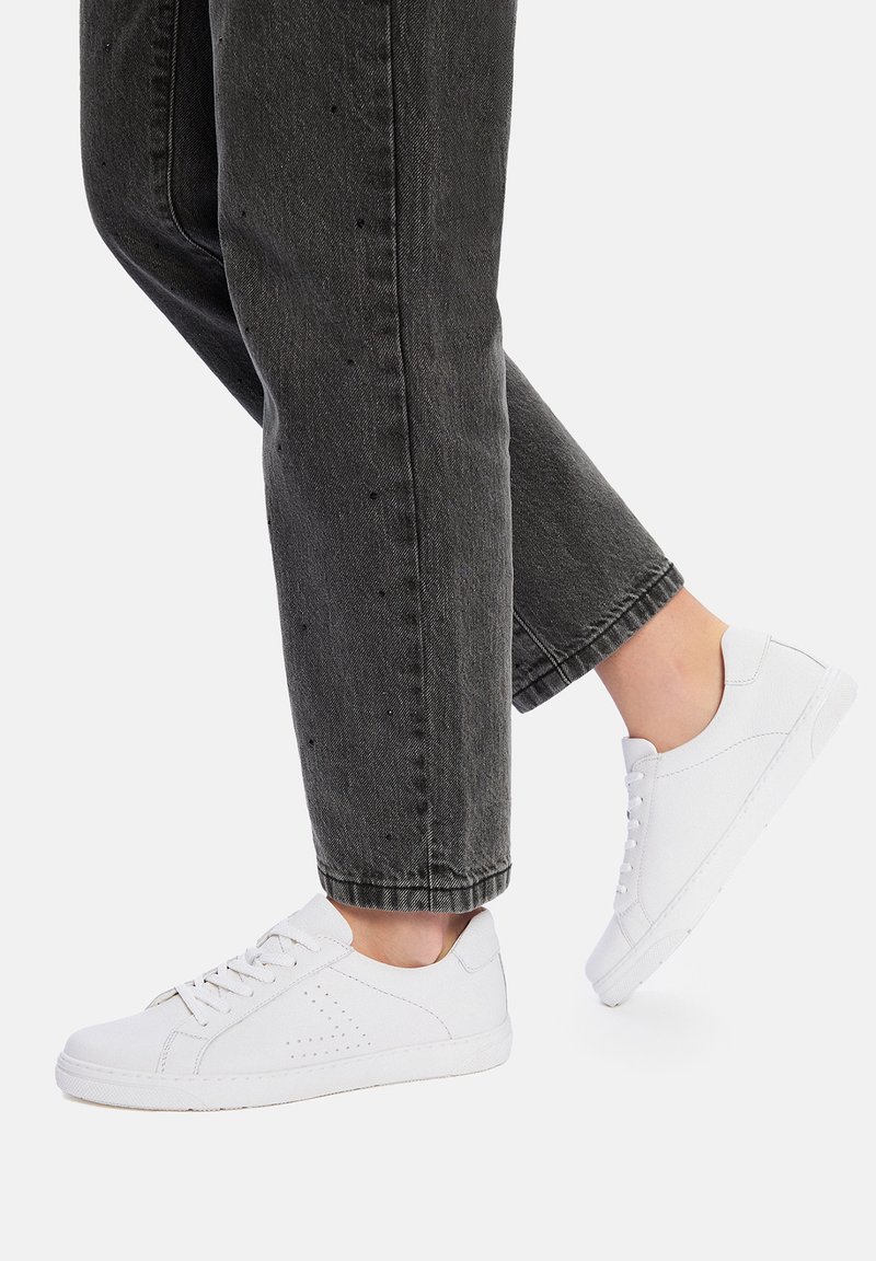 Białe sneakersy z perforowanymi akcentami, sznurowane, z gumową podeszwą, zestawione z przyciętymi czarnymi dżinsami. Prosta i czysta estetyka.