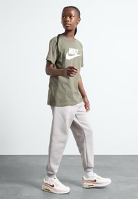 T-shirt en coton vert olive avec logo Nike blanc, associé à un pantalon de survêtement gris clair et des baskets blanches avec des accents roses et rouges.