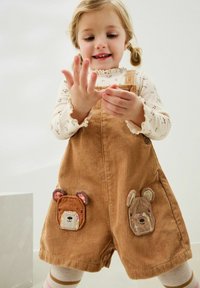 Gli overall in corduroy marrone presentano toppe a forma di faccia d'orso su ciascuna tasca. Sotto, viene indossata una maglietta a maniche lunghe di colore crema con un motivo floreale e maniche a volant.