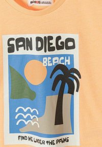 Diseño de camiseta con «San Diego Beach», un sol, una colina verde, una palmera, olas del océano y el texto «Find me under the palms» en tela color durazno.