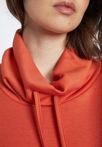 Oranges Sweatshirt mit Kragen und Bindebändern. Glatte Textur, leichtes Material, Nahaufnahme zeigt den Kragen- und Halsbereich.